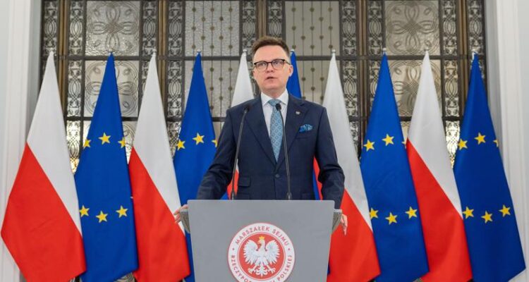 Marszałek Sejmu Szymon Hołownia stojący na tle flag Polski oraz Unii Europejskiej.