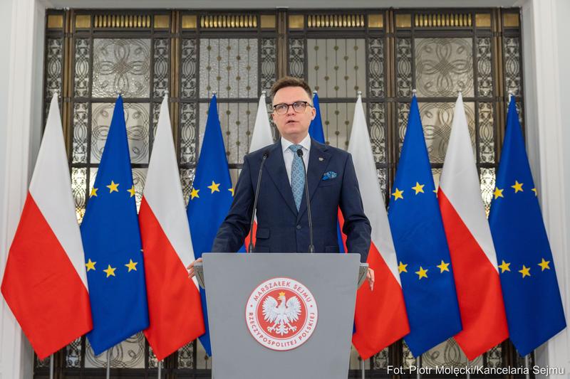 Marszałek Sejmu Szymon Hołownia stojący na tle flag Polski oraz Unii Europejskiej.